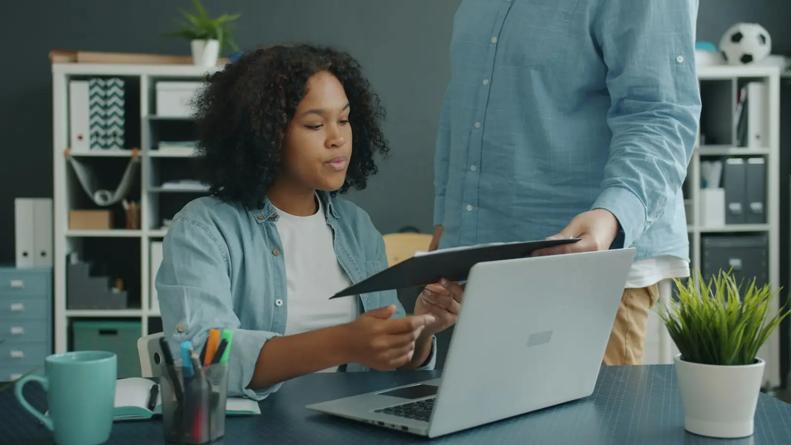 A colleague handing documents to a woman working at a laptop